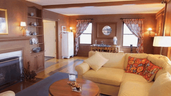 A warm-toned room features a sofa, a decorated shelf, and sunlight filtering through patterned curtains.