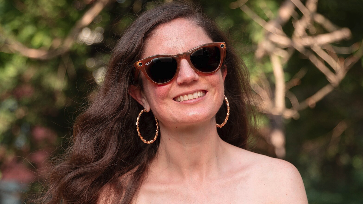 Woman outdoors smiling, wearing sunglasses and hoop earrings, surrounded by blurred green foliage.
