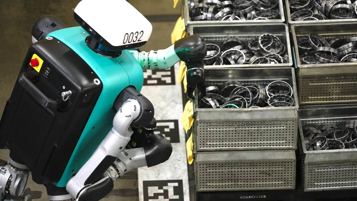 A humanoid robot with teal and white elements handles metal parts in bins on a factory floor.