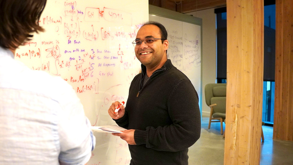 Tanmay Gupta is pictured smiling next to a whiteboard filled with mathematical formulas, embodying active AI engagement.