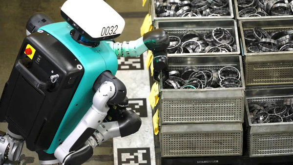 A humanoid robot with teal and white elements handles metal parts in bins on a factory floor.