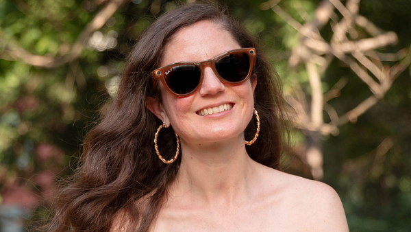 Woman outdoors smiling, wearing sunglasses and hoop earrings, surrounded by blurred green foliage.
