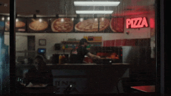Through a rainy window, a pizza worker prepares food beneath menu boards and a red neon "Pizza" sign.
